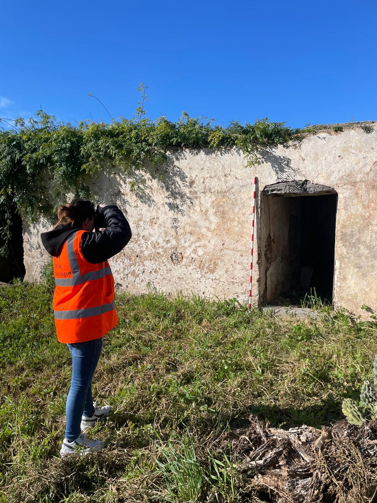 Arqueóloga con chaleco naranja fotografía una estructura de piedra antigua junto a una vara topográfica.
