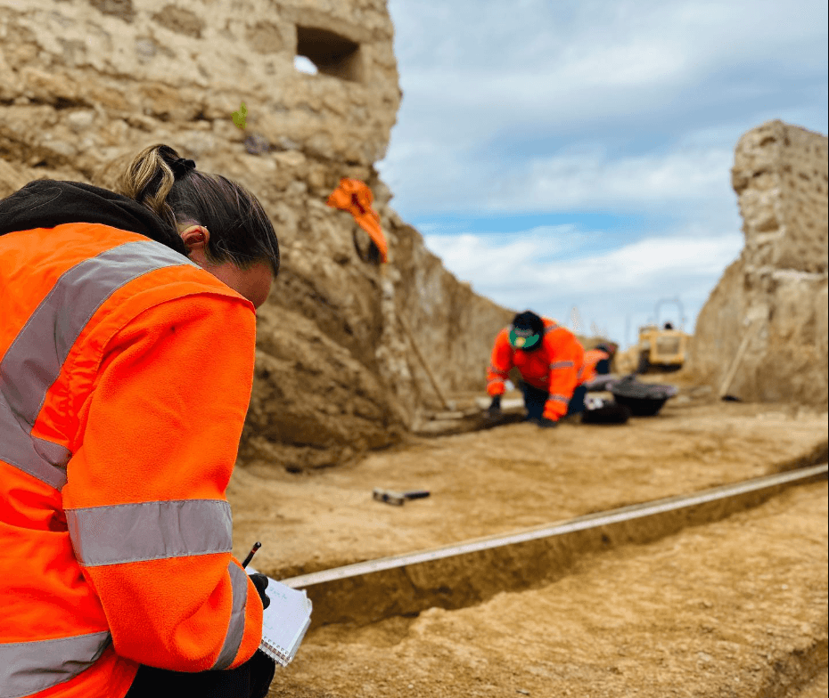 Arqueóloga toma notas mientras un colega trabaja en una excavación cerca de ruinas antiguas.