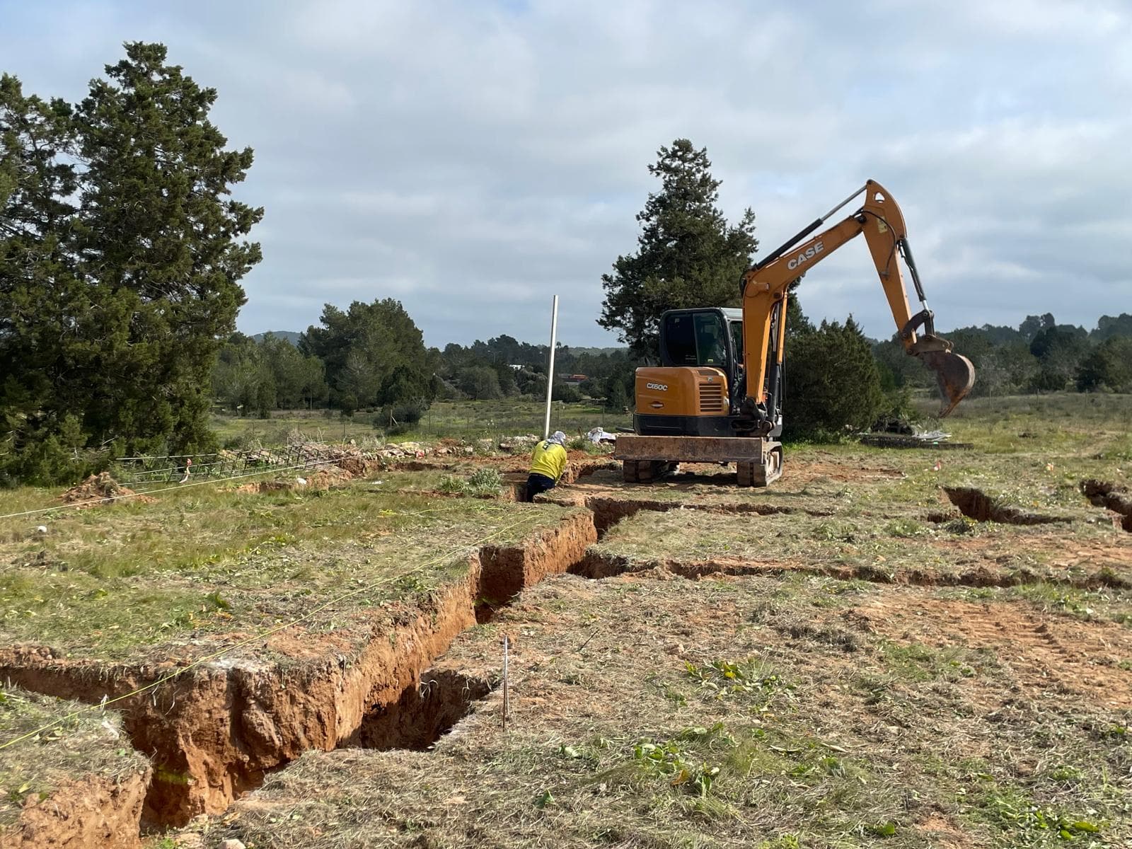 Excavadora naranja cavando zanjas en un terreno rural con un trabajador y árboles al fondo.