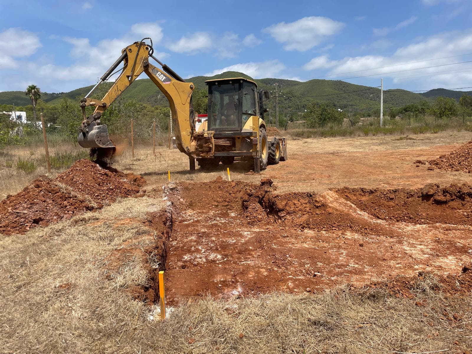 Retroexcavadora amarilla excavando tierra rojiza en un campo abierto bajo un cielo azul despejado.
